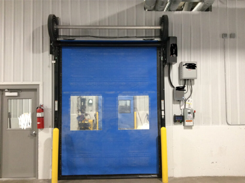 Large commercial blue roll up garage door with two windows next to electric panels hanging on wall